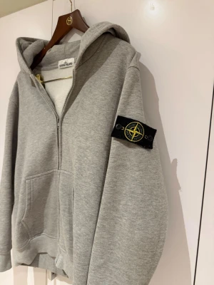  Stone Island Zip Hoodie - Snygg grå hoodie med dragkedja från Stone Island. Klassisk modell med huva, känguruficka och ikonisk logga på vänster ärm. Mjuk bomullskvalitet som är perfekt för en avslappnad stil.