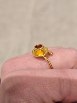 Guldfärgad ring med tekoppdesign - Unik guldfärgad ring formad som en liten tekopp med fat. Ringen har en röd sten i mitten av koppen som ger en cool detalj. Perfekt för dig som gillar annorlunda och lekfulla accessoarer. Materialet är metall med guldfinish.