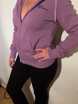 Lila hoodie från Peak Performance - Säljer en lila hoodie från Peak Performance med dragkedja framtill och två fickor. Hoodien har en stor huva och ribbade muddar vid ärmslut och nederkant. Perfekt för chill dagar eller när du vill ha en avslappnad stil.