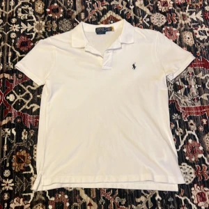 Vit pikétröja från Ralph Lauren - Klassisk vit pikétröja från Ralph Lauren med korta ärmar och den ikoniska broderade loggan på bröstet. Tröjan har krage och knappslå framtill, tillverkad i mjuk bomull för en bekväm känsla. Går gärna ner i pris vid snabb affär