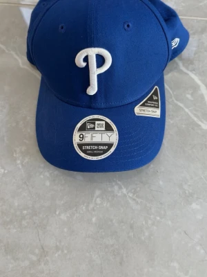 Blå New Era 9Fifty keps - Snygg blå keps från New Era, modell 9Fifty Stretch-Snap. Framtill sitter ett vitt broderat P och kepsen har klassisk böjd skärm. Insidan har svart svettband och detaljer med basebollmotiv. Perfekt för dig som gillar sportig stil.