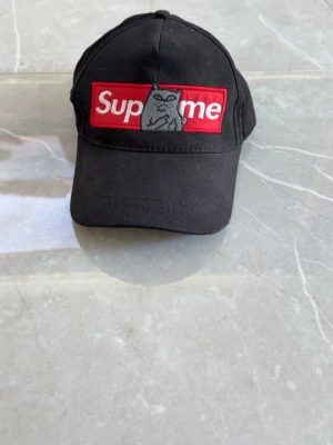 Svart Supreme keps med kattmotiv - Säljer en svart keps från Supreme med klassisk röd box logo framtill och ett broderat kattmotiv i mitten av loggan. Kepsen har böjd skärm och justerbar passform. Tillverkad i bomullstyg som känns både slitstarkt och bekvämt.