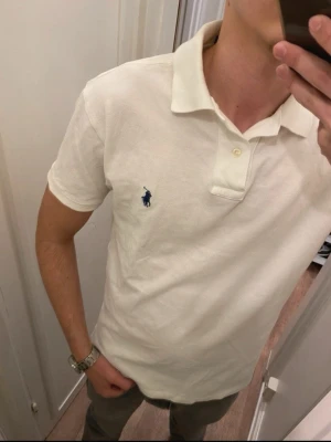 Vit pikétröja från Polo Ralph Lauren - Klassisk vit pikétröja från Polo Ralph Lauren med blå broderad logga på bröstet. Tröjan har krage, korta ärmar och knappar vid halsen. Tillverkad i mjuk bomull för en bekväm känsla och passar perfekt till jeans eller chinos.