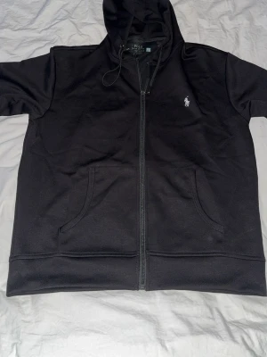 Svart hoodie från Polo Ralph Lauren - Snygg svart hoodie från Polo Ralph Lauren med dragkedja framtill och klassisk vit logga broderad på bröstet. Hoodien har två stora fickor fram och justerbar huva med snören. Tillverkad i mjukt bomullsmaterial som känns skönt mot huden.