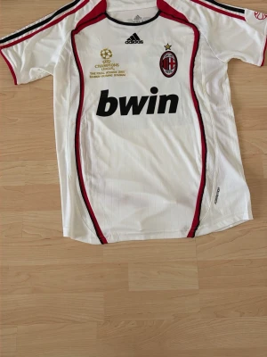 AC Milan vit matchtröja Adidas - Säljer en vit AC Milan matchtröja från Adidas med svarta och röda detaljer. Tröjan har klubbmärke, Champions League-detaljer och Bwin som sponsortryck på bröstet. Tillverkad i lätt och ventilerande material, perfekt för fotbollsträning eller match.