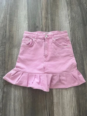 Rosa volangkjol med fickor - Söt rosa kortkjol med volangkant nertill och klassiska jeansfickor fram och bak. Kjolen har bälteshällor och stängs med knapp och dragkedja. Perfekt för dig som vill ha en lekfull och trendig look.