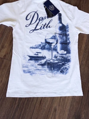 Cain & Marcel T-Shirt - Vit t-shirt med ett stort blått tryck på ryggen som föreställer en vinflaska, ett vinglas och en båt vid kusten. T-shirten har klassisk passform och rund hals. Perfekt för dig som gillar unika motiv och stilrena färger.