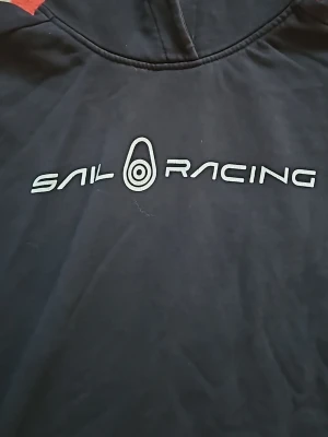 Svart hoodie från Sail Racing - Snygg svart hoodie från Sail Racing med logga tryckt i vitt på bröstet. Klassisk huva och avslappnad passform, perfekt för en sportig och casual stil. Materialet känns mjukt och bekvämt mot huden.