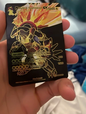 Pokémonkort M Hybrid Typhlosion EX - Unikt Pokémonkort med M Hybrid Typhlosion EX-motiv. Kortet har svart bakgrund med guld- och rödfärgade detaljer och holografisk effekt. Text och illustrationer är i metallic guld. Perfekt för samlare eller Pokémonfans.