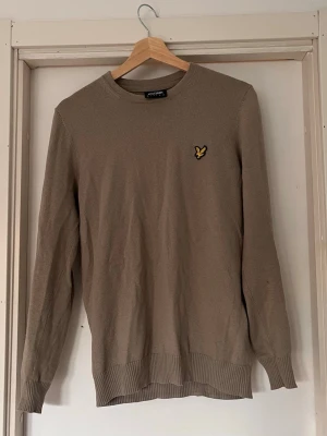 Beige stickad tröja Lyle & Scott - Snygg beige stickad tröja från Lyle & Scott med rund halsringning och den klassiska gula loggan på bröstet. Tröjan har långa ärmar och ribbade muddar vid ärmslut och nederkant. Perfekt för lager på lager och enkel att matcha. Tröjan är i storleken XS.