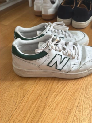 Vita New Balance sneakers med gröna detaljer - Säljer ett par klassiska vita New Balance sneakers med gröna detaljer och stor N-logga på sidan. Skorna har lågt skaft, vita snören och är tillverkade i läder. Perfekta för dig som gillar retrostil och vill ha något som funkar till det mesta.