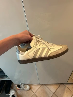 Adidas Samba vita sneakers - Klassiska Adidas Samba sneakers i vitt med gråa detaljer och bruna gummisulor. Skorna har de ikoniska tre ränderna på sidan och texten 'SAMBA' i guld. Ovandelen är i läder och mocka, vilket ger en retro och sportig vibe.