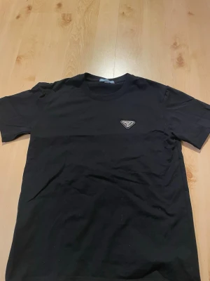 Svart t-shirt från Prada - Svart t-shirt från Prada med klassisk passform och rund hals. Diskret Prada-logga på bröstet. Tillverkad i mjuk bomull som känns skön mot huden. Perfekt för dig som gillar stilrena och exklusiva plagg.