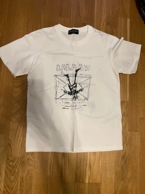 Vit Balmain t-shirt med tryck - Snygg vit t-shirt från Balmain med svart grafiskt tryck framtill. Klassisk passform med rund hals och korta ärmar. Tillverkad i mjuk bomull som känns skön mot huden. Perfekt för dig som gillar streetstyle och exklusiva märken.