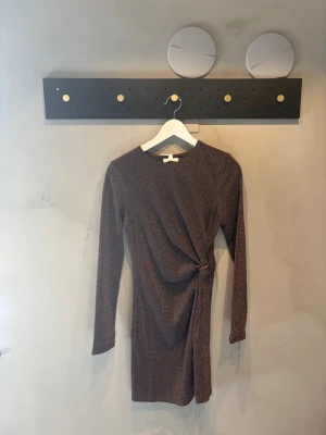 Glittrig brun kortklänning - Kort klänning i brun ton med glittrigt tyg och lång ärm. Klänningen har en snygg drapering och twistdetalj i midjan som ger en cool siluett. Perfekt för dig som vill sticka ut lite extra. Ingen synlig logga på märket.