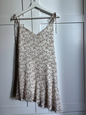 Blommig kortklänning med knytband - Söt kortklänning i ljusbeige med små blommor i rosa toner. Klänningen har smala axelband med knyt, v-ringad hals och snedskuren nedtill 