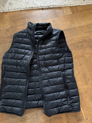 Svart dunväst Peak Performance - Snygg svart dunväst från Peak Performance med quiltad design och hög krage. Västen är ärmlös och har dragkedja framtill samt två praktiska fickor med dragkedja. Perfekt att slänga över en hoodie eller tröja för extra värme.