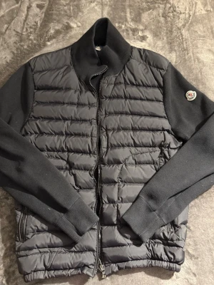 Svart Moncler Cardigan - Snygg svart Moncler Cardigan från Moncler med quiltad front och rygg, samt stickade ärmar och hög krage. Jackan har dragkedja framtill och Moncler-logga på ena ärmen. Perfekt för kalla dagar och riktigt trendig stil.  Inga hål eller så använd ett par gånger dustbag medföljer 