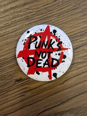 Punks Not Dead pinsknapp - Cool knapp med texten 'Punks Not Dead' i svart graffiti-stil. 4,4 cm. Perfekt accessoar för dig som gillar punk och vill sticka ut. Rund form och klassisk punkkänsla.