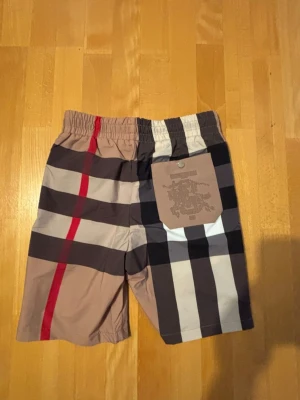Nya Burberry shorts - Snygga burberryshorts med klassiskt rutigt mönster i beige, brunt, svart, vitt, blått och rött. Shortsen har elastisk midja med svart snörning och sidofickor. Perfekta för sommaren och ger en trendig look.