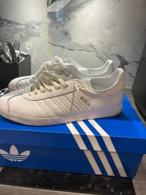 Vita Adidas Gazelle sneakers Herr - Snygga vita Adidas Gazelle sneakers i läder med klassiska tre ränder på sidan och guldfärgad Gazelle-text. Skorna har vit sula och vita skosnören, perfekt för en clean och stilren look. Ikonisk modell som funkar till det mesta.