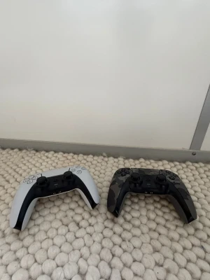 Stick drift fråga om du inte vet vad de ärPS5 DualSense handkontroller svart & vit - Två PlayStation 5 DualSense handkontroller, en i klassiskt vit och svart design och en i svart med grå kamouflagemönster. Ergonomisk form med greppvänliga handtag och responsiva knappar. Tillverkade i slitstark plast, perfekta för gaming på PS5.