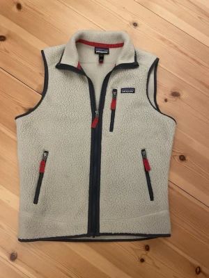 Äkta beige fleeceväst från Patagonia - Mysig beige fleeceväst från Patagonia med mörkblå detaljer och röda dragkedjor. Västen har tre fickor med dragkedja och hög krage. Perfekt för att ha under tishan på varma dagar i sommar och enkel att matcha med olika outfits. Endast använd ett fåtal gånger så den är i princip ny. Hör av er vid frågor!