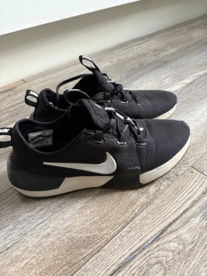 Svarta Nike sneakers med vit sula - Snygga svarta Nike sneakers med vit sula och vit swoosh på sidan. Ovandelen är i meshmaterial för bra andningsförmåga och skorna har svarta snören samt dragflikar med Nike-logga. Perfekta för en sportig och avslappnad stil. Storlek 40,5