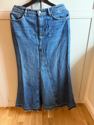 Lång jeanskjol i blå denim - Säljer en lång jeanskjol i klassisk blå denim med femficksmodell och rå kant nertill. Kjolen har hög midja, bälteshällor och en snygg slits framtill för extra rörelsefrihet. Perfekt till sneakers eller boots för en cool vardagslook.