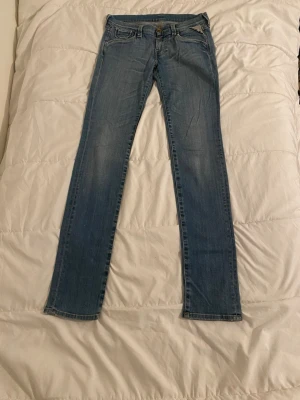 Blå raka jeans från Replay - Säljer ett par klassiska blå jeans från Replay med raka ben och låg midja. Jeansen har snygga slitningar, fem fickor och detaljer i bronsfärgad metall. Materialet är mjukt denim i bomull med kontrastsömmar och Replay-logga på knappen.