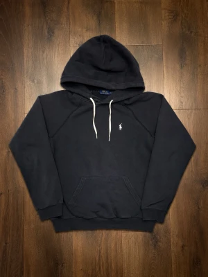 Mörkblå hoodie från Polo Ralph Lauren - Snygg mörkblå hoodie från Polo Ralph Lauren med vit broderad logga på bröstet och vita dragsnören i huvan. Ett litet hål vid magen som är jätte enkelt att sy ihop men syns knappt 