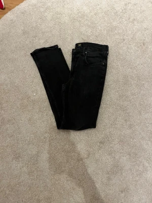 Svarta slim fit jeans från Lee - Säljer ett par svarta slim fit jeans från Lee med klassisk femficksdesign och knappgylf. Jeansen har raka ben och är tillverkade i stretchigt denimtyg för extra komfort. Perfekta till en avslappnad streetwear-look.