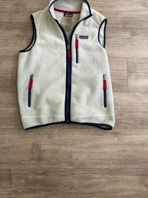 Patagonia Väst - Säljer en väldigt populär beige fleeceväst från Patagonia med marinblå detaljer och röda dragkedjor. Västen har tre fickor med dragkedja och står för en sportig och avslappnad stil. Perfekt att slänga på sig över en hoodie eller tröja.