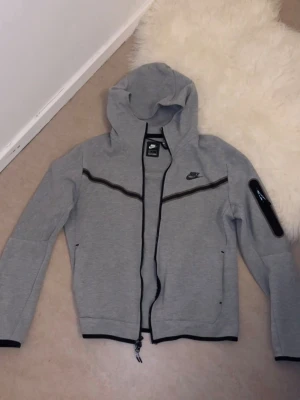 Grå Nike Tech Fleece hoodie - Snygg grå Nike Tech Fleece hoodie med svart dragkedja och detaljer. Har huva, fickor med dragkedja och en extra ficka på ärmen. Perfekt för en sportig och chill look. Tillverkad i mjukt och bekvämt material.