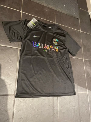 Svart Balmain x Nike t-shirt - Snygg svart t-shirt från Balmain x Nike med regnbågsfärgad logga och PSG-märke på bröstet. Tillverkad i ett glansigt syntetmaterial som känns sportigt och modernt. Klassisk rund hals och korta ärmar, perfekt för en trendig streetwear-look.