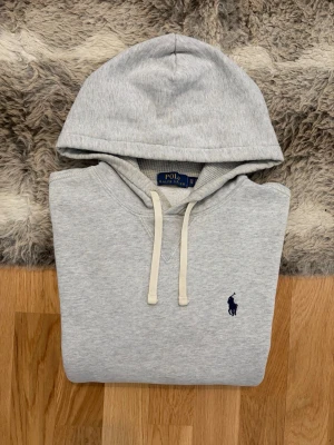 Ljusgrå Polo Ralph Lauren Hoodie - Hej! 👋 Säljer en skön och klassisk ljusgrå Polo Ralph Lauren-hoodie i storlek XS. Den har den stilrena mörkblå loggan broderad på bröstet 🐎 Tröjan är i ganska gott skick, men jag vill vara helt tydlig med att den har visst slitage på ärmsluten samt lite på axeln (se bilder för detaljer). Det är inget som stör helhetsintrycket vid användning, men priset är satt därefter! ✨ 100% äkta med tvättlappar kvar. Perfekt som en vardagströja där komforten ligger i fokus. Fraktar snabbt! 🚛📦💬💯 