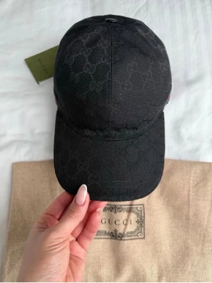 Svart keps från Gucci med GG-mönster - Snygg svart keps från Gucci med diskret GG-monogram över hela kepsen. På sidan finns ett klassiskt grönt och rött band. Kepsen är tillverkad i canvasmaterial och har böjd skärm. Perfekt accessoar för en trendig och exklusiv look.