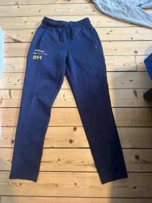 Marinblå mjukisbyxor från CCM - Säljer ett par marinblå mjukisbyxor från CCM med broderade detaljer och dragkedjefickor. Byxorna har resår och snörning i midjan samt raka ben. Perfekta för chill eller träning. Materialet känns mjukt och bekvämt mot huden.
