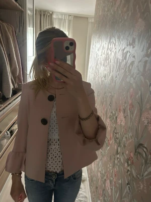 Ljusrosa kappa med volangärm - Trendig ljusrosa kappa med stora svarta knappar och volangdetaljer vid ärmsluten. Kortare modell som ger en modern siluett. Finns en fläck, bild finns, men det kan vara så att den går bort i tvätten
