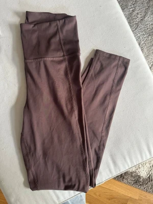 Bruna träningstights från Kay/Day - Snygga bruna träningstights från Kay/Day i storlek S. De har hög midja och är gjorda i ett mjukt och stretchigt material som sitter skönt på kroppen. Perfekta för att matcha med oversized hoodies eller crop tops. Stitchade detaljer längs benen för en clean look.