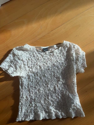 Vit spetsblus från Gina Tricot - Säljer en söt vit blus från Gina Tricot med korta ärmar och ett genombrutet spetsmönster. Toppen har en figurnära passform och är perfekt för dig som gillar romantiska detaljer och vill ha något extra till din outfit.