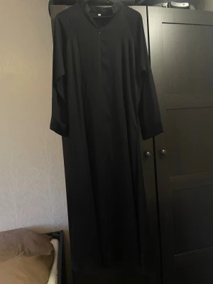 Svart Silky Abaya - Saudiarabien - Svart abaya som är silky i storlek 52. Använd få gånger och är som ny. Köpt från Saudi Arabien. Abayan kan bäras som stängd modell, eller öppen, men då behöver man justera den nerifrån så att dragkedjan går hela vägen ner.  Längd: ca 132 cm. Ärmlängd: ca 66 cm. Passar de som är under 160 cm, skulle jag säga.