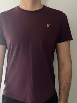 Vinröd t-shirt från Lyle & Scott - Snygg vinröd t-shirt från Lyle & Scott med klassisk gul logga på bröstet. Tillverkad i mjuk bomull och har en normal passform med korta ärmar och rund hals. Perfekt till jeans eller shorts för en clean look. Storlek L men ENBART PASSFORM FÖR M
