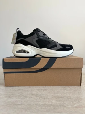 Svarta och grå sneakers från Skechers - Stilrena sneakers från Skechers i svart och grått med vita sulor och synlig luftdämpning i hälen. Ovandelen är i mesh och syntetmaterial med snygga detaljer och logga på sidan. Perfekta för en sportig och trendig look.
