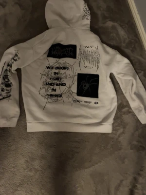 Vit hoodie med svart tryck och dragkedja - Säljer en vit hoodie med huva och dragkedja framtill. Hoodien har flera svarta grafiska tryck på både fram- och baksida samt på ärmarna, inklusive taggar, spindel och text. Stora fickor framtill och broderad text över magen. Perfekt för dig som gillar streetwear och unik design.