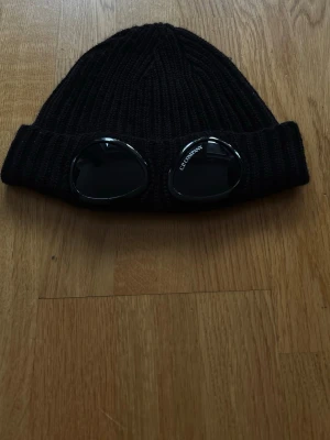 Svart goggle-mössa från C.P. Company - Säljer en svart ribbstickad mössa från C.P. Company med de ikoniska goggle-linserna framtill. Mössan är i grovstickad ullmix och har en bred uppvikt kant. Perfekt för dig som vill ha streetwear-vibe och unik stil.