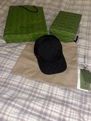 Svart keps från Gucci med monogram - Snygg svart keps från Gucci med klassiskt GG-monogram över hela kepsen. På sidan finns en ikonisk grön och röd rand. Kepsen är i tygmaterial och har justerbar rem bak. Perfekt för dig som vill ha en exklusiv och trendig accessoar.