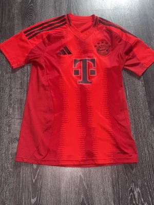 Röd Bayern München tröja Adidas S - Säljer en röd Bayern München matchtröja från Adidas med svarta detaljer och klubbmärke på bröstet. Tröjan har korta ärmar, V-ringning och tre svarta ränder på axlarna. Tillverkad i lätt och ventilerande material, perfekt för fotbollsträning eller match.