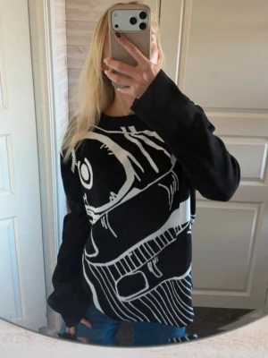 Svart stickad tröja med grafiskt tryck - Cool svart stickad tröja med ett stort vitt grafiskt tryck av en hand som håller en sprayburk. Tröjan har rund halsringning och långa ärmar. Perfekt för dig som gillar streetwear och unika plagg.