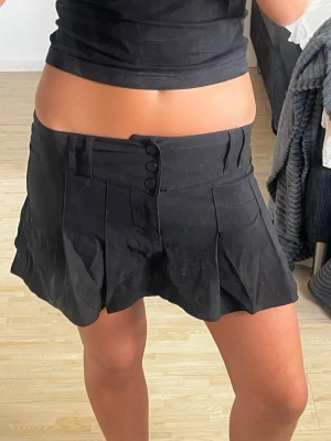 Svart plisserad minikjol med knappar - Säljer en svart kortkjol med plisserad design och dekorativa knappar framtill. Kjolen har hög midja och en lätt utsvängd passform som ger en cool och trendig look. Perfekt att matcha med crop tops eller hoodies. Midjemått: 35cm (stretchiga). Längd: 29 cm